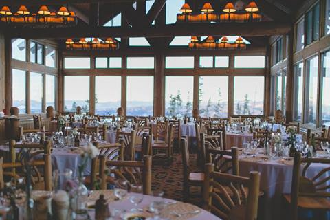 Wedding reception in a wood lodge with chandeliers, multiple set tables, and floor to ceiling windows. 