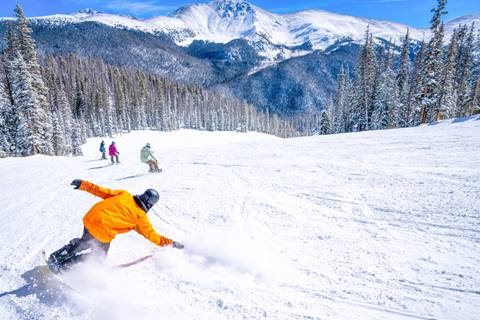 Snowboarders skiing down a groomed run with mountains in the background at Winter Park Resort