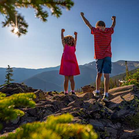 Two children raise their arms up in joy and celebration as they stand at the summit of Winter Park near Sunspot Lodge