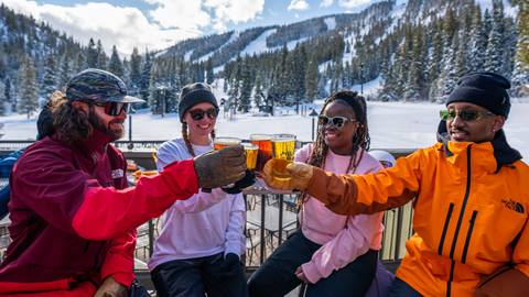 Four friends enjoying après at Mary Jane at Winter Park Resort, toasting with drinks on a sunny outdoor deck with a snowy mountain backdrop.