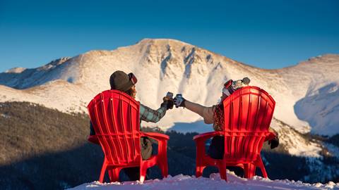 two people sitting in red chairs cheersing beers in the winter on top of a mountain