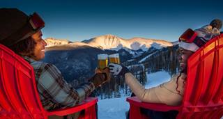 Two people sitting in red chairs outdoor at dusk with the sun setting on mountains in the background